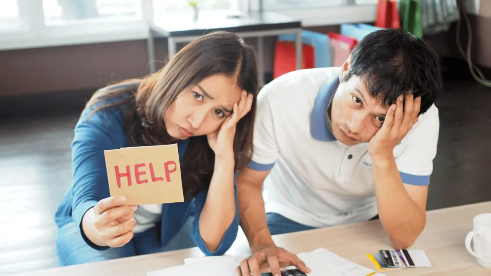 Stressed couple at a laptop holding a sign that says HELP while searching for financial assistance